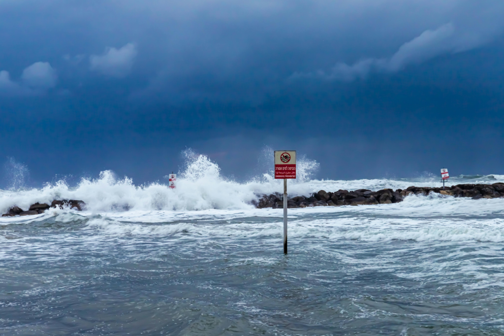 Waves crash against rocks beneath dark storm clouds near a no swimming warning sign.