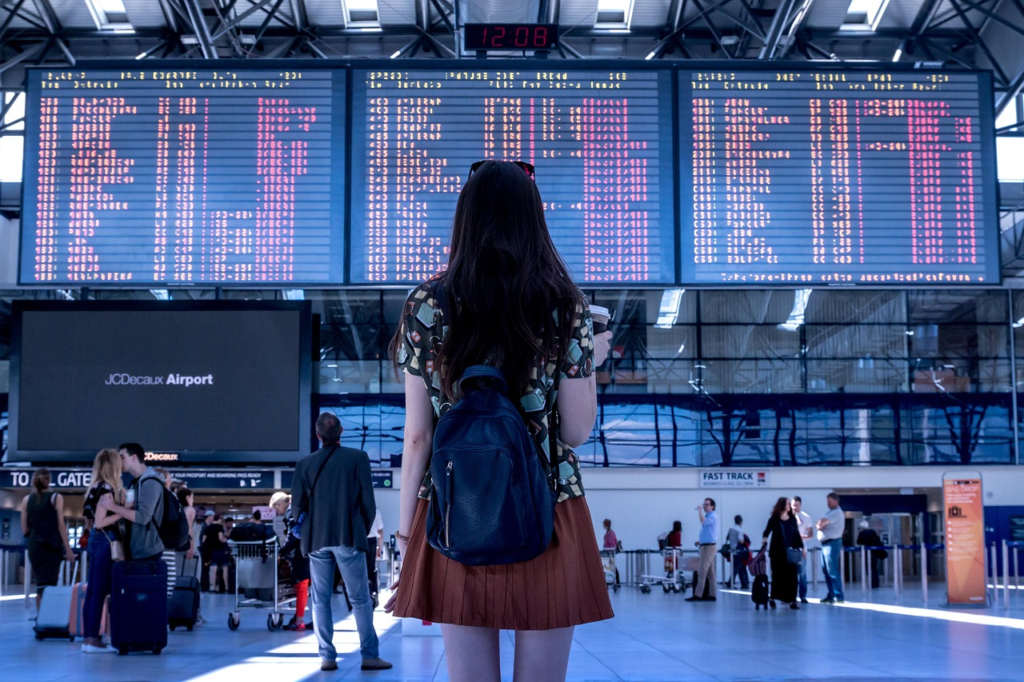 Woman with a backpack looking at a large airport departure board inside a busy terminal.