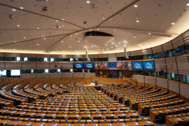 Interior of the European Parliament plenary chamber with curved rows of desks and EU flags.