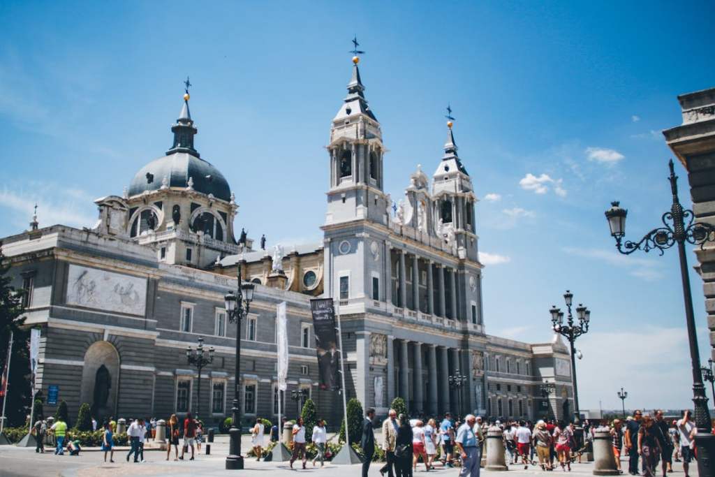 People walk in a busy plaza outside a historic cathedral with tall towers and a domed roof under a clear sky.