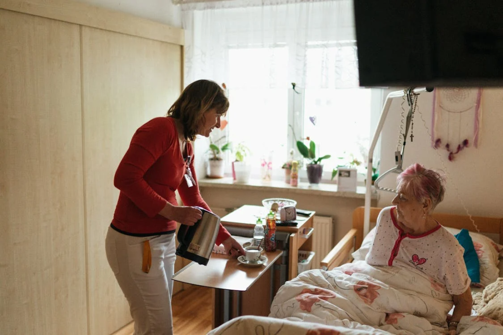 Caregiver pours a drink for an elderly woman sitting in bed inside a bright, home-style care room.