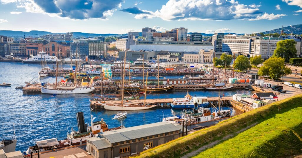 Oslo harbor with boats docked, a city skyline in the background, and a sunny sky with scattered clouds.
