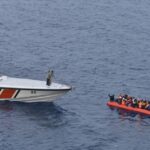 Turkish Coast Guard boat approaches crowded inflatable raft carrying migrants at sea.