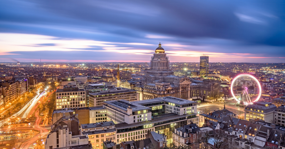 Brussels cityscape at dusk with illuminated buildings, a domed palace, and a glowing Ferris wheel on the skyline.