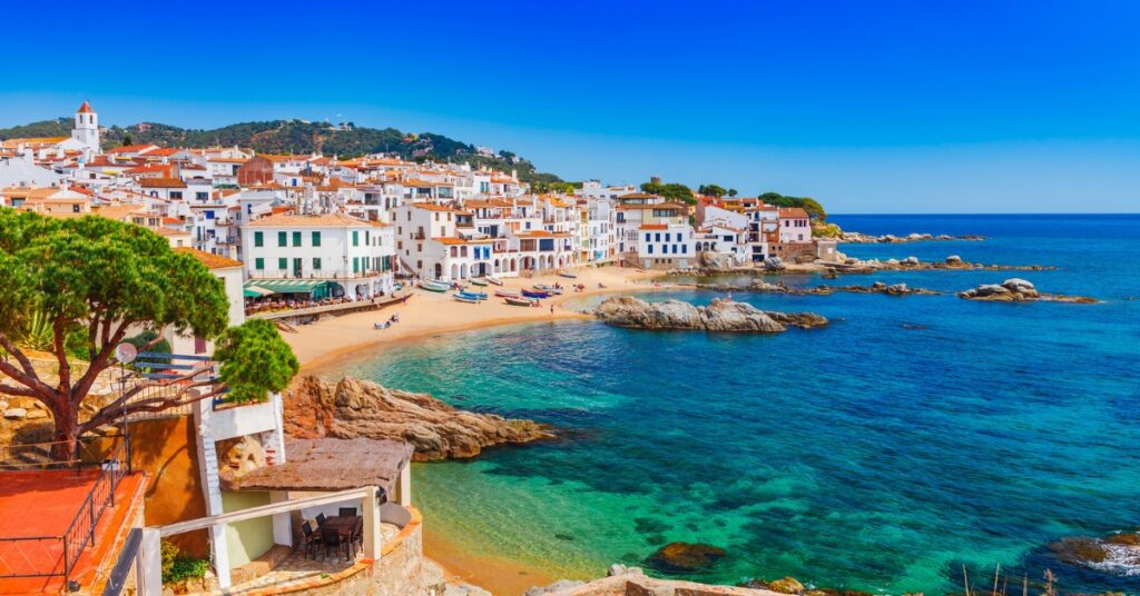 Coastal town with white buildings and red roofs beside a beach and turquoise sea under a clear blue sky.
