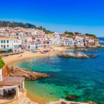 Coastal town with white buildings and red roofs beside a beach and turquoise sea under a clear blue sky.