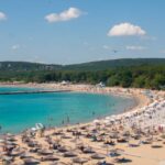 Crowded beach with rows of umbrellas and people swimming in clear blue water along the shoreline
