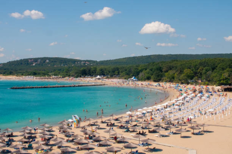 Crowded beach with rows of umbrellas and people swimming in clear blue water along the shoreline