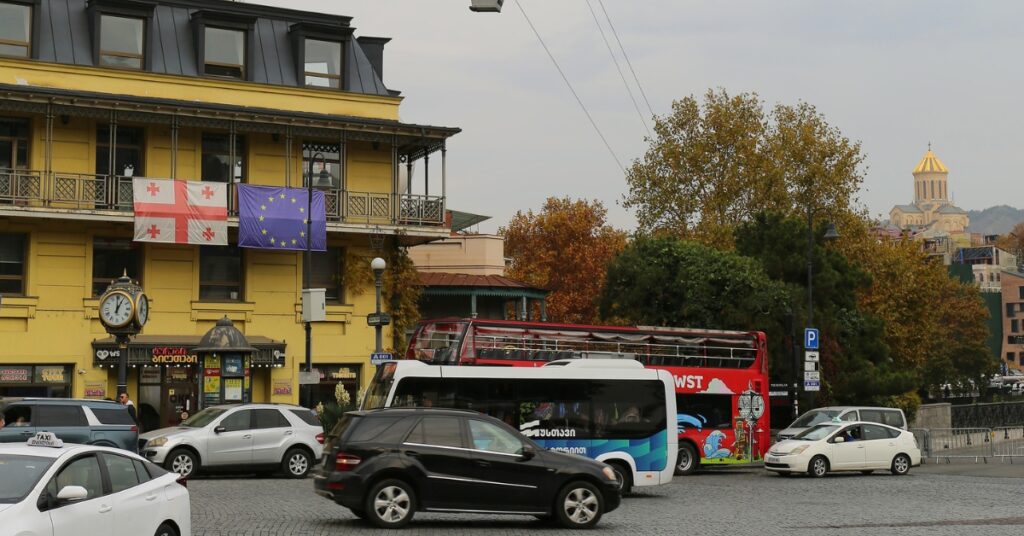Georgia and European Union flags displayed on a city building above a busy street with cars and buses.