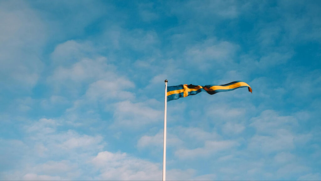 Swedish flag waving on a tall flagpole against a blue sky with scattered clouds.