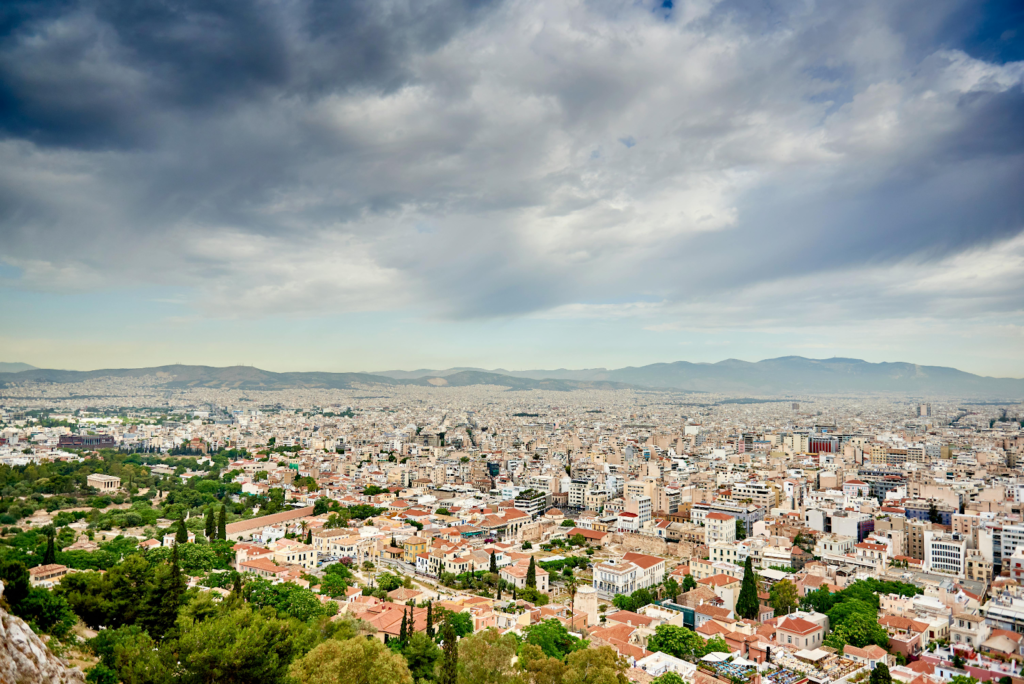 Panoramic view of Athens, Greece with dense city buildings, distant mountains, and dramatic clouds overhead.