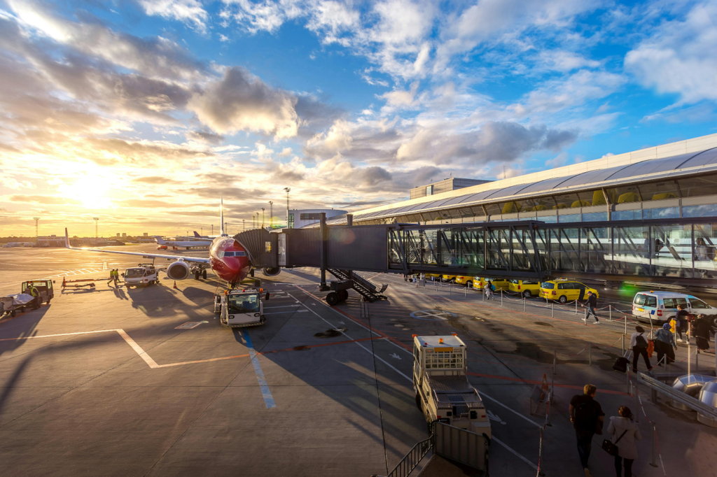 Commercial airplane parked at an airport gate connected to a jet bridge with ground vehicles nearby at sunset.
