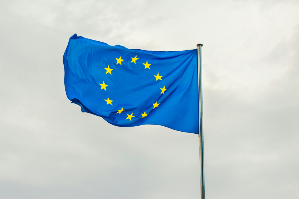 European Union flag with yellow stars waving on a flagpole against a cloudy sky.