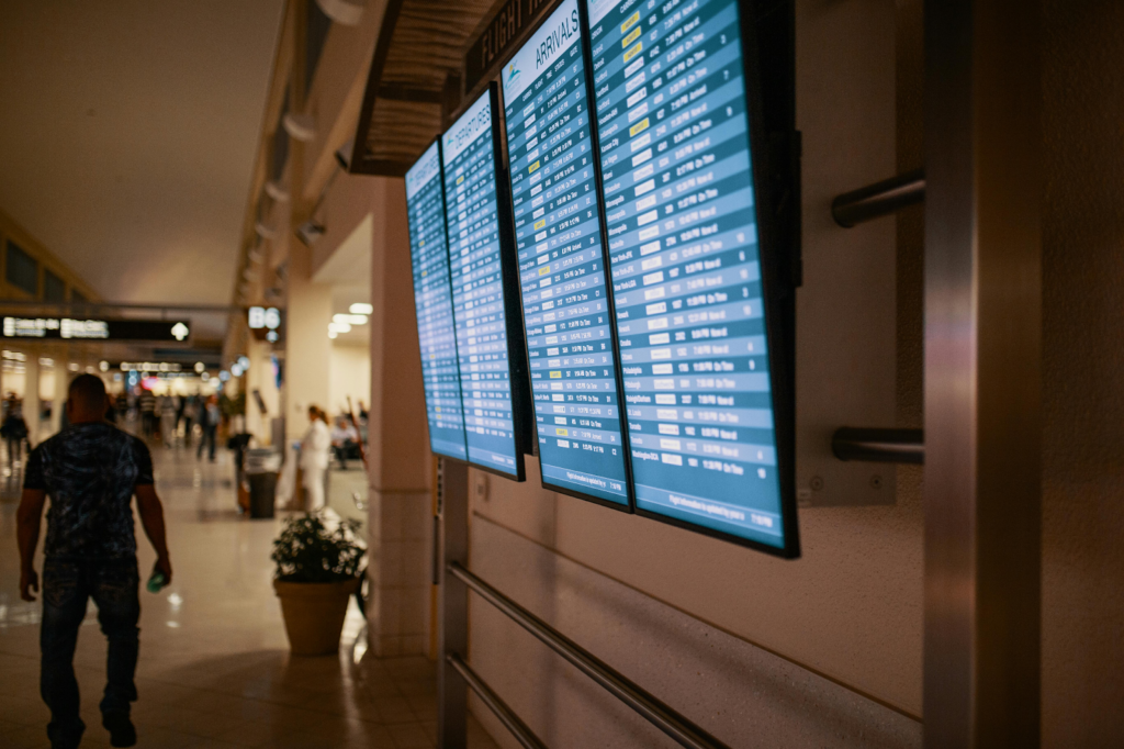 Digital airport arrival boards displaying flight information inside a busy terminal corridor.