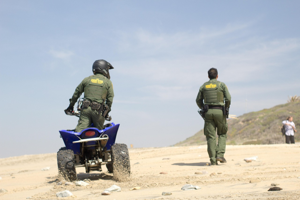 Two border patrol agents patrol a sandy beach, one riding an ATV while the other walks ahead