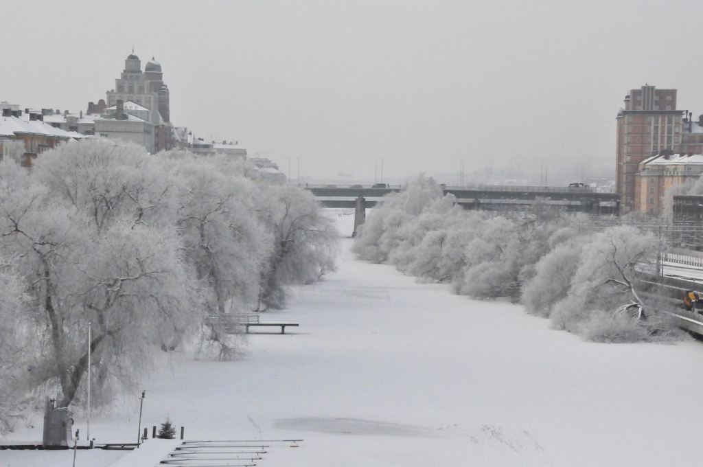 Snow-covered river lined with frost-covered trees and city buildings in winter.