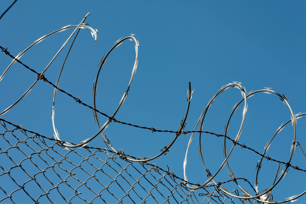 Razor wire coils and barbed wire along the top of a chain-link fence against a clear blue sky