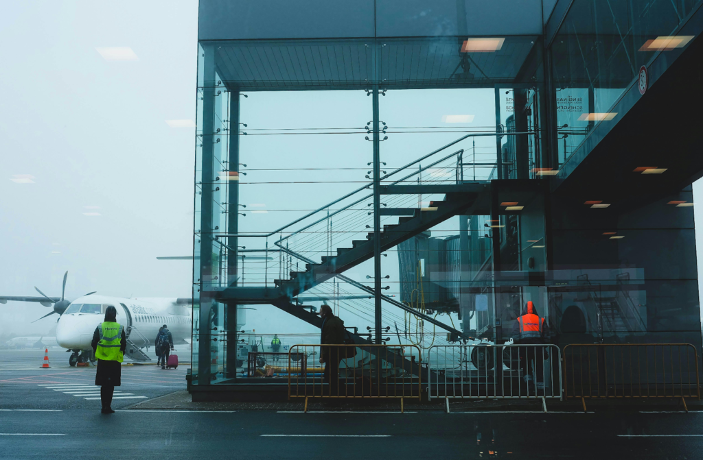 Glass airport terminal with stairs, workers in vests, and airplane on a foggy tarmac.
