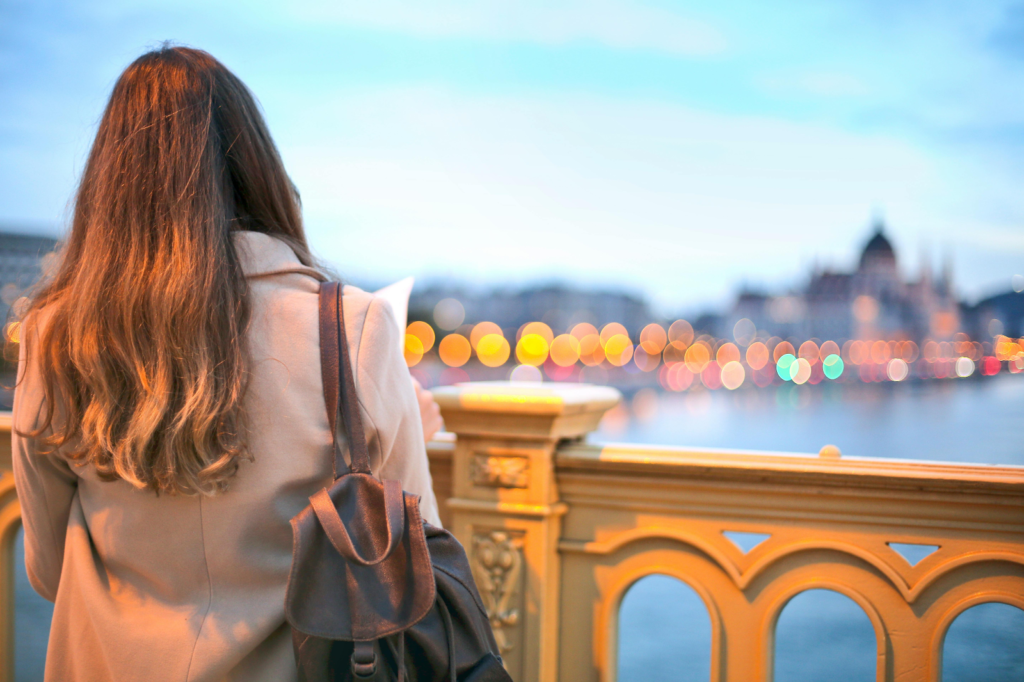 Woman with backpack stands on a bridge overlooking a river and city lights at dusk.