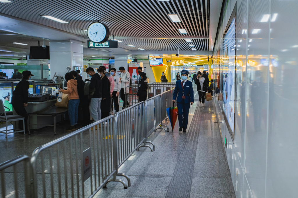 Passengers line up at an airport security check while a uniformed staff member walks through the terminal.