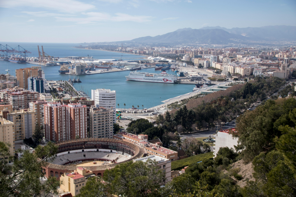 Panoramic view of a coastal city harbor with ships, urban skyline, and mountains in the background.