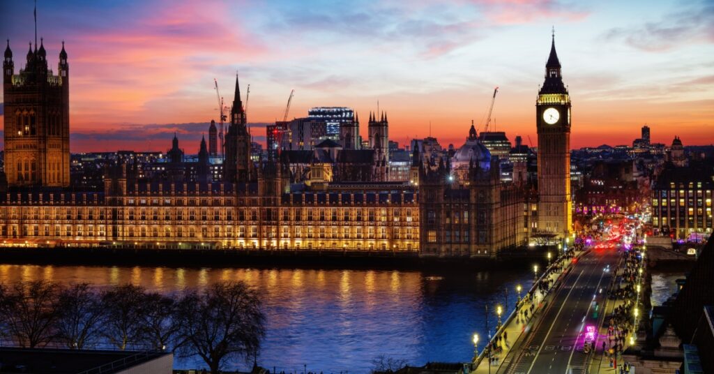 Sunset view of Big Ben and the Houses of Parliament with a lit bridge over the Thames River.