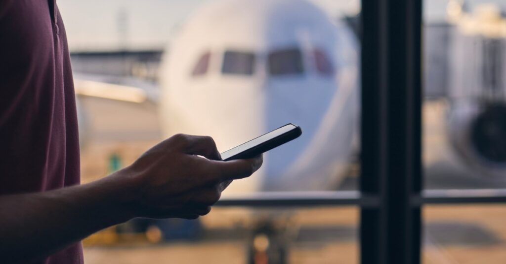 Person using a smartphone near an airport window with a parked airplane visible outside