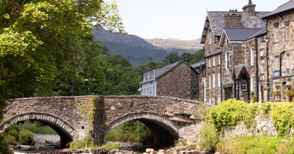 Stone bridge over a shallow river beside rustic village houses with trees and mountains in the background.