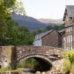 Stone bridge over a shallow river beside rustic village houses with trees and mountains in the background.