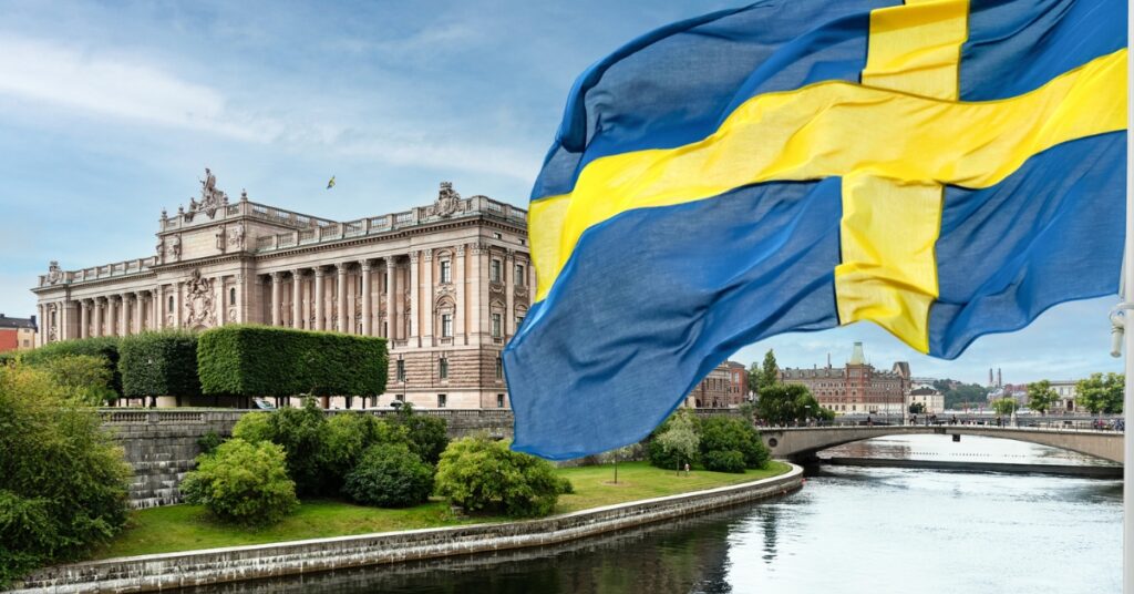 Swedish flag waving above a river with the Swedish Parliament building and bridge in Stockholm in the background.