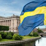 Swedish flag waving above a river with the Swedish Parliament building and bridge in Stockholm in the background.