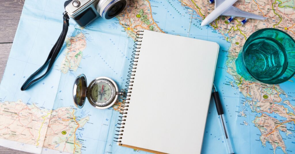 Travel flat lay with map, camera, notebook, compass, toy airplane, and glass on a table.