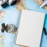 Travel flat lay with map, camera, notebook, compass, toy airplane, and glass on a table.