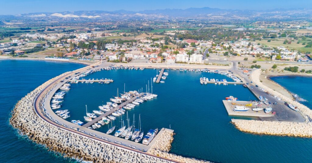 Aerial view of a marina with boats inside a curved breakwater beside a coastal town and mountains.