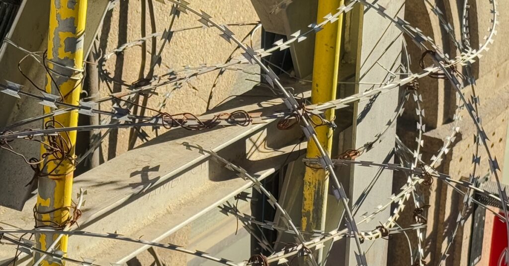 Coiled barbed wire wrapped around a metal fence with yellow posts, casting sharp shadows in sunlight
