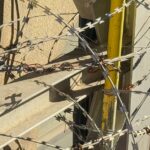 Coiled barbed wire wrapped around a metal fence with yellow posts, casting sharp shadows in sunlight