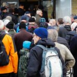 Crowd of people queued outside a building entrance, many wearing jackets and backpacks