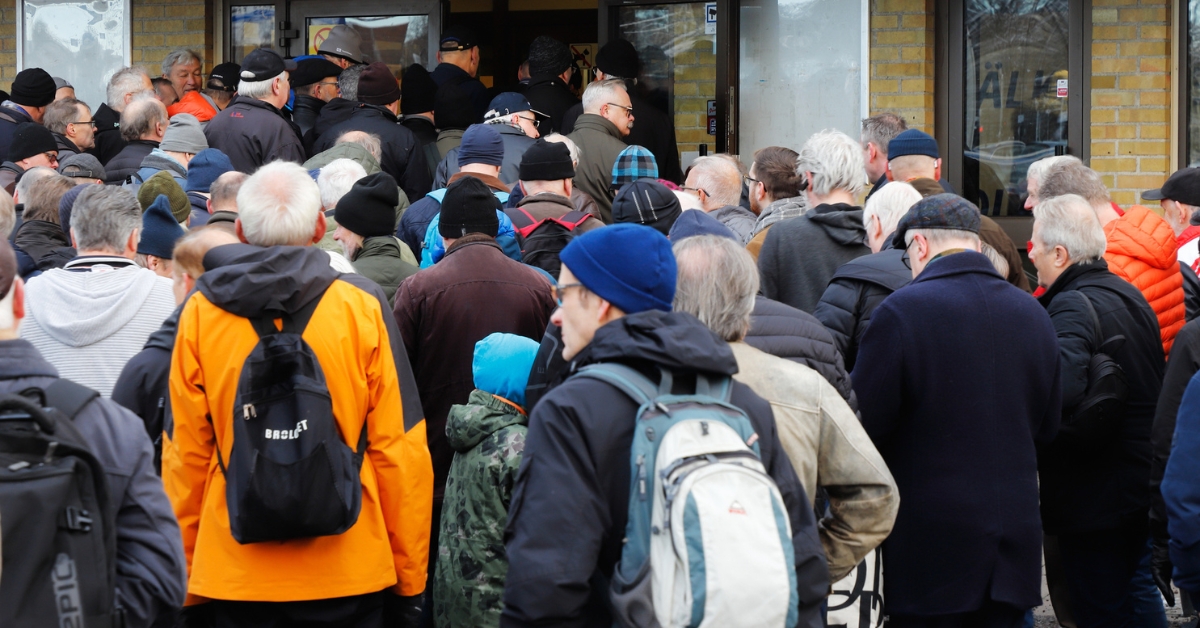 Crowd of people queued outside a building entrance, many wearing jackets and backpacks