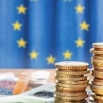 Stacks of euro coins and banknotes beside a calculator with the EU flag in the background.
