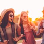Four friends wearing backpacks and sunglasses walk together outdoors at sunset, smiling and talking