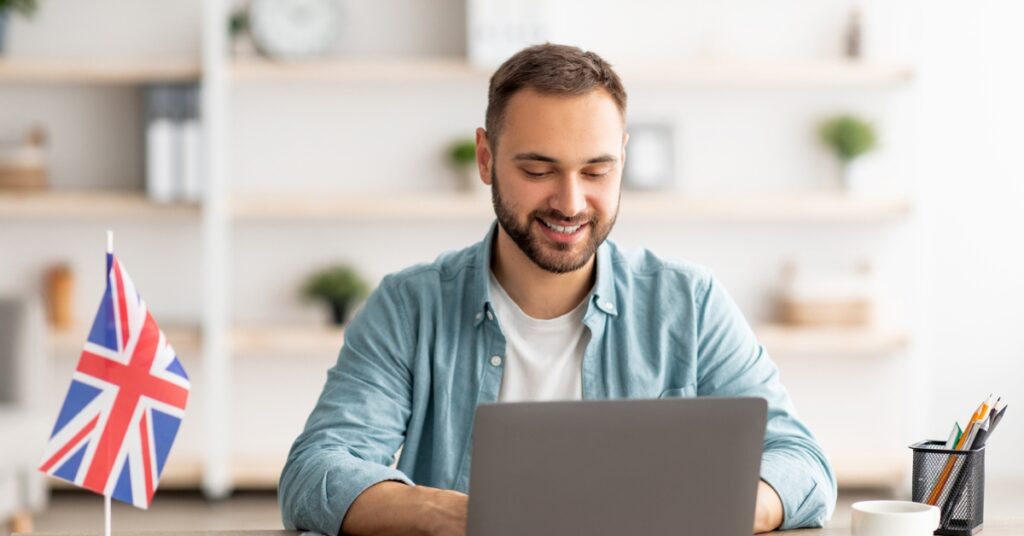 Man smiling while using a laptop at a desk with a small UK flag, suggesting online English learning