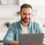Man smiling while using a laptop at a desk with a small UK flag, suggesting online English learning