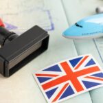 Passport with stamps, rubber stamp, toy airplane, and UK flag sticker on travel documents.