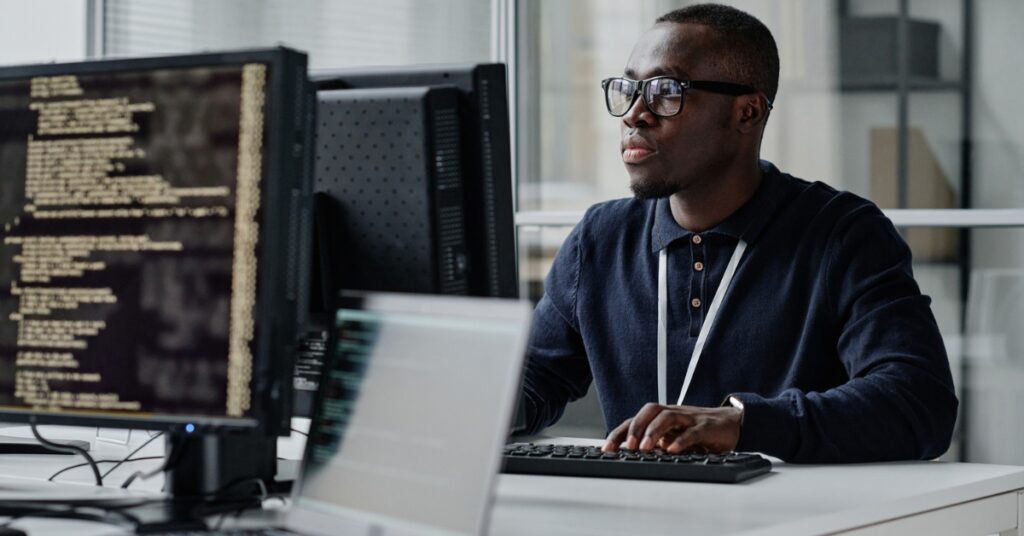 Software developer working at a computer with multiple monitors displaying code in an office setting