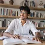 Student smiling while studying with books and a laptop in a library setting.