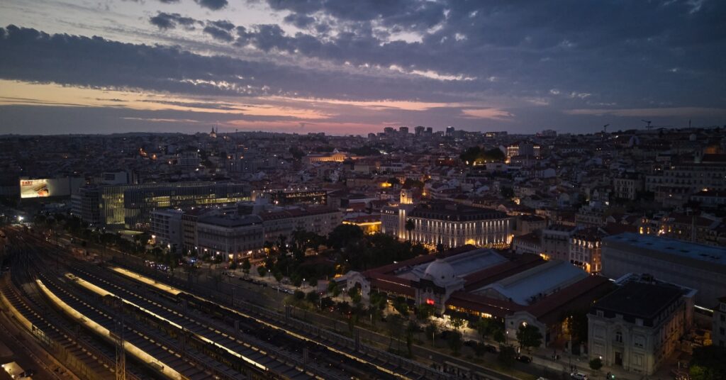 City skyline at dusk with illuminated buildings and train tracks beneath a cloudy evening sky.