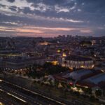 City skyline at dusk with illuminated buildings and train tracks beneath a cloudy evening sky.