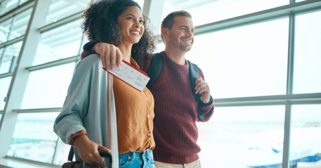 Smiling couple walks through an airport terminal holding luggage and a boarding pass.