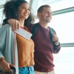 Smiling couple walks through an airport terminal holding luggage and a boarding pass.