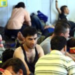 People rest on the floor inside a crowded shelter while others sit and drink water.